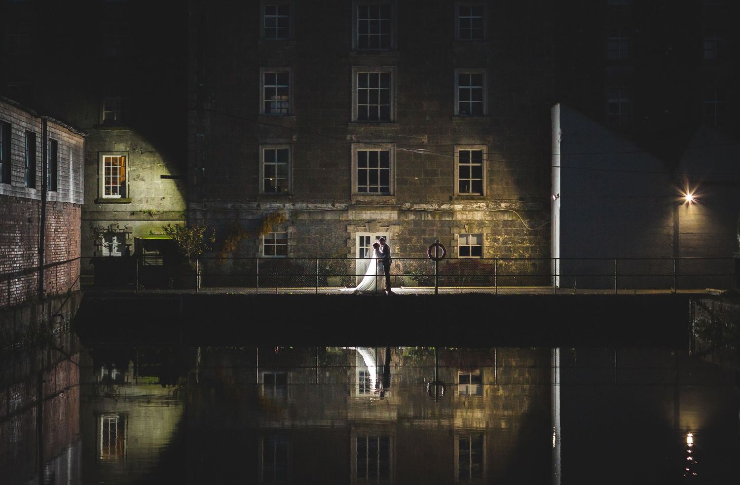 A nighttime reflection of figures on a bridge over dark canal waters with illuminated buildings.