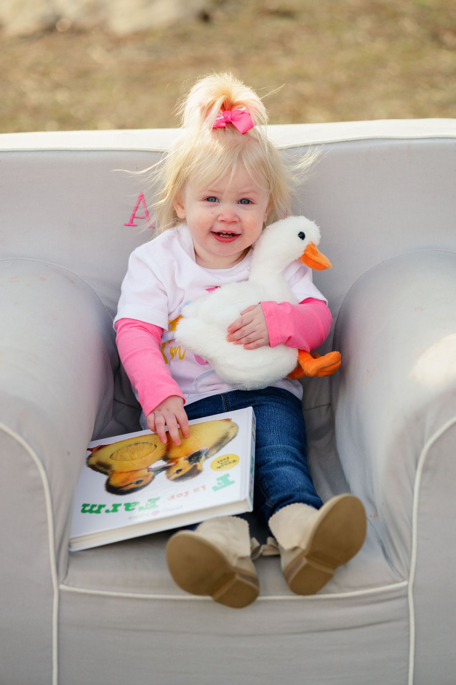 A child sits in a white armchair reading a farm book while holding a plush duck toy.