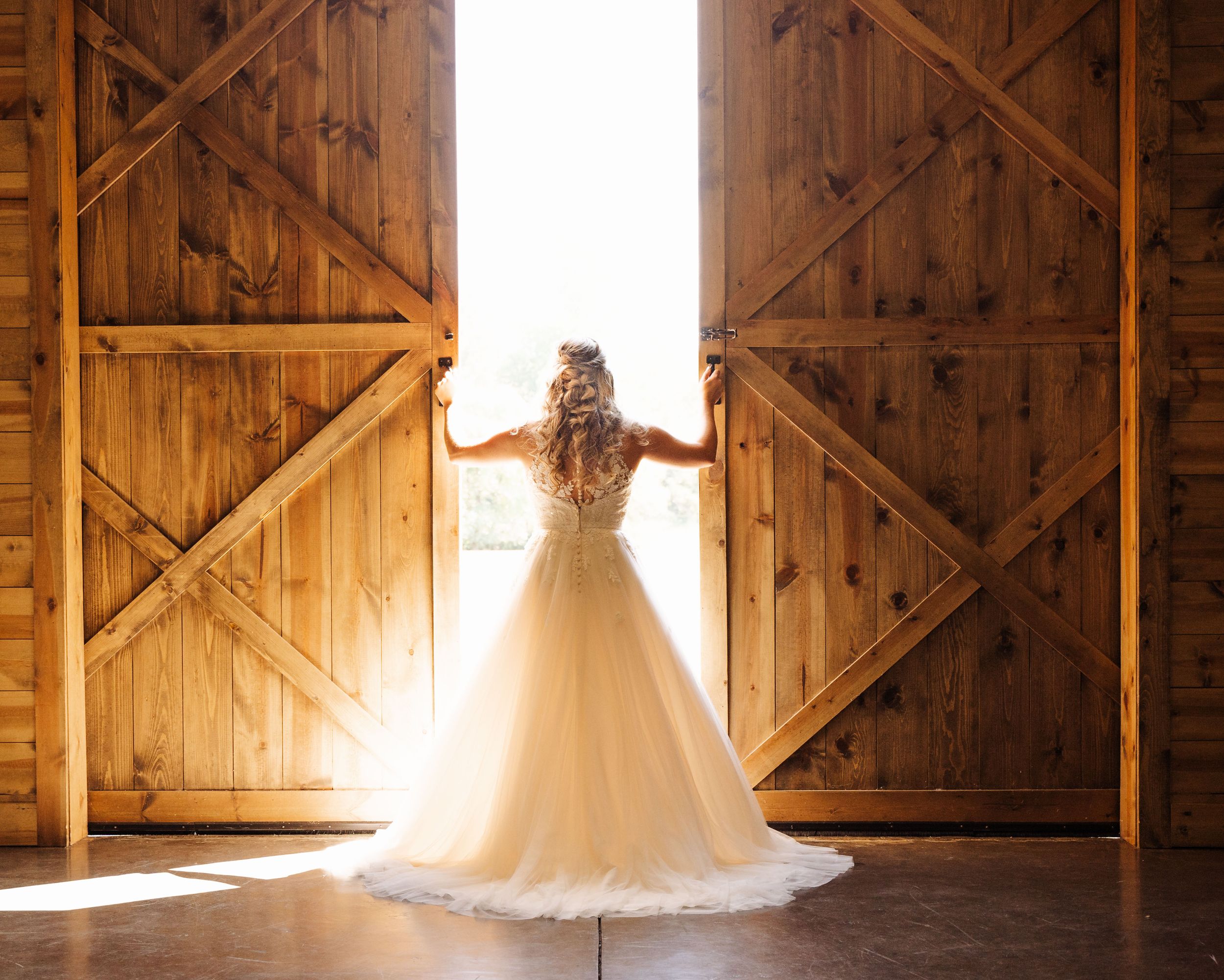 Bride poses in front of barn doors with sun streaming through her dress at Hidden Acres in Gillsville, Georgia
