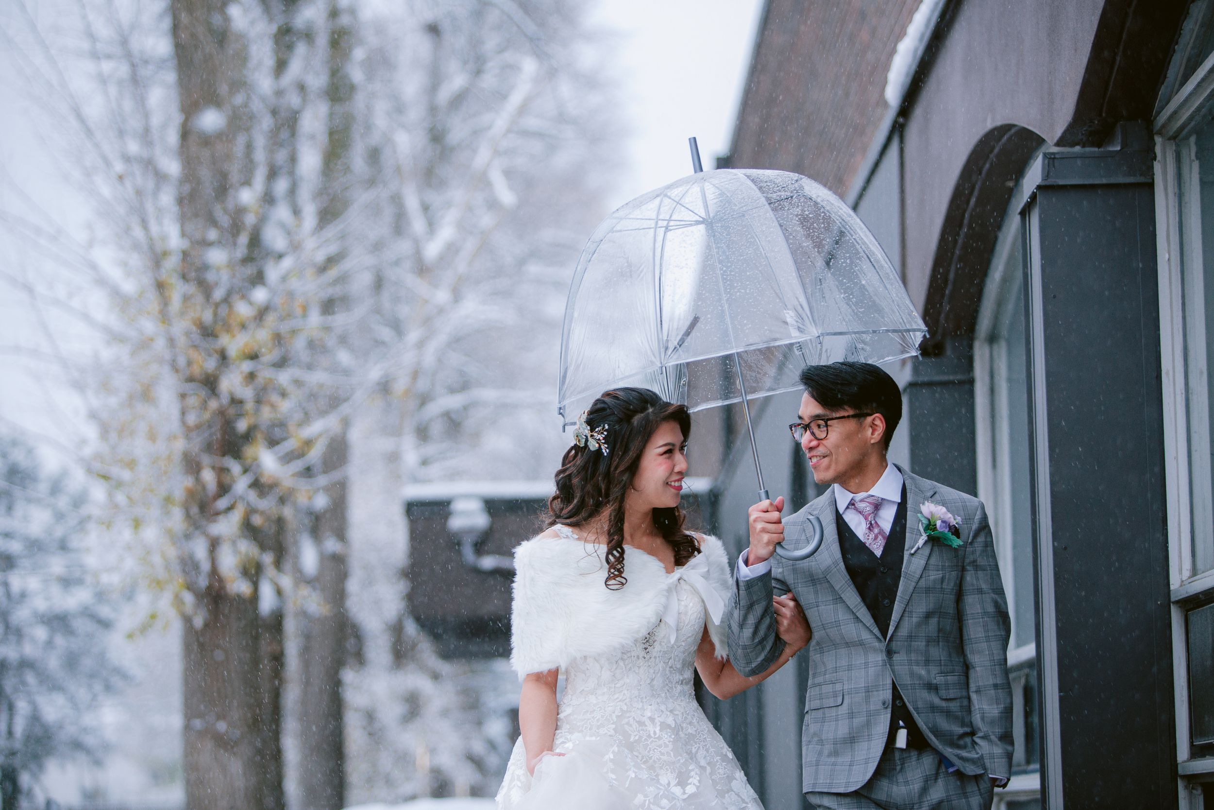 A couple shares an umbrella during their winter wedding photoshoot in a snowy forest setting.