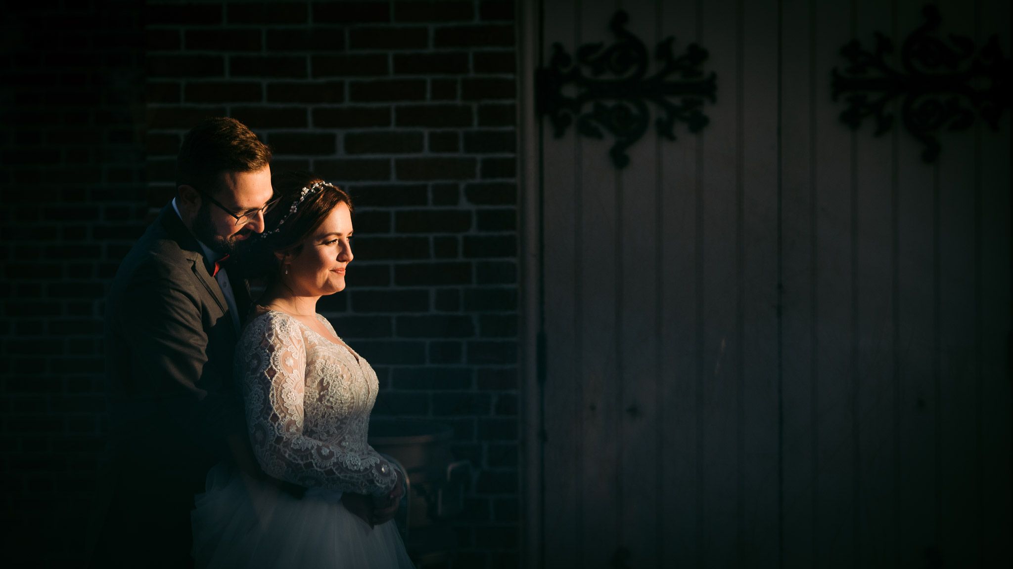 Dramatic moody portrait against brick wall with light highlighting lace wedding dress details.
