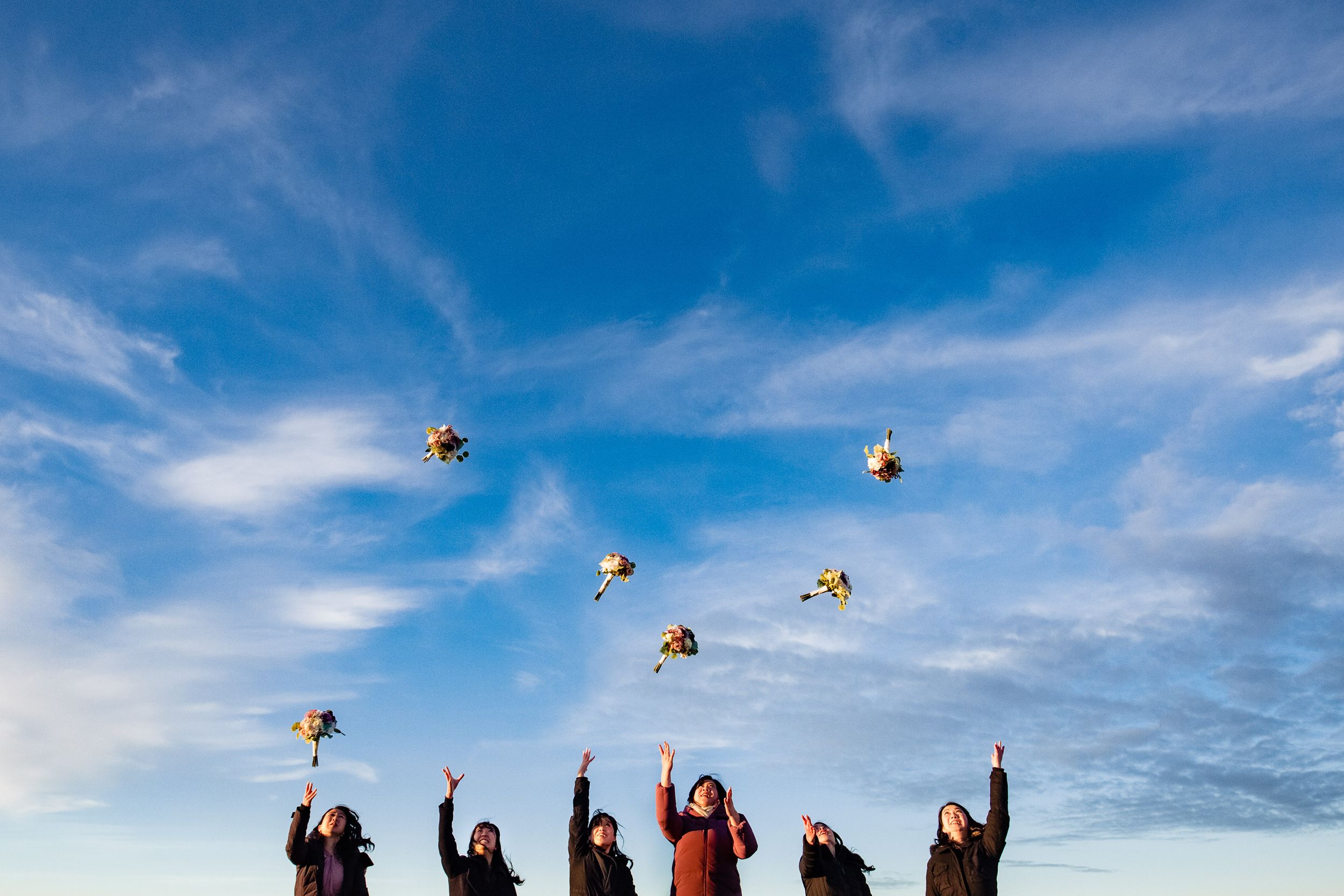 Wedding party throwing bouquets into bright blue sky during celebration.