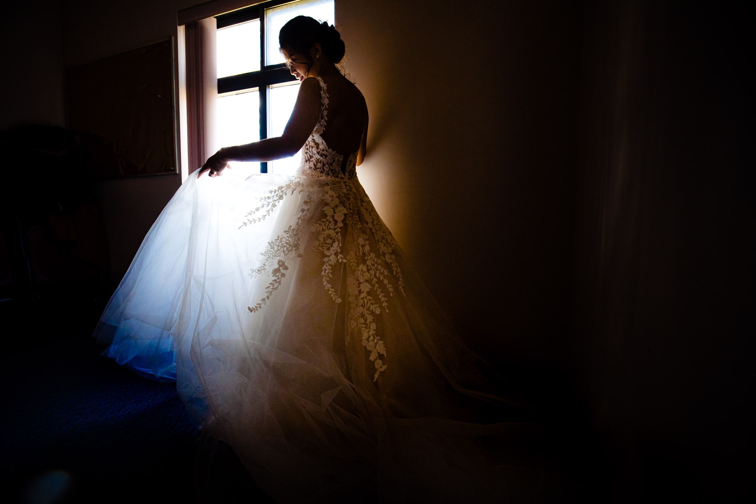 Silhouette of a bride in a flowing ballgown dress standing by a window in dramatic lighting.