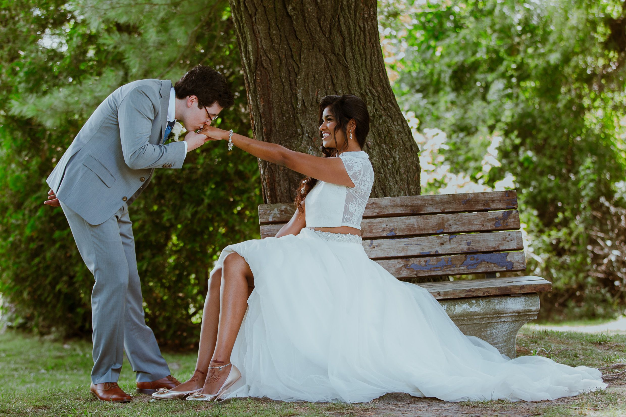 Groom kisses bride's hand while she sits on rustic wooden bench in garden setting.