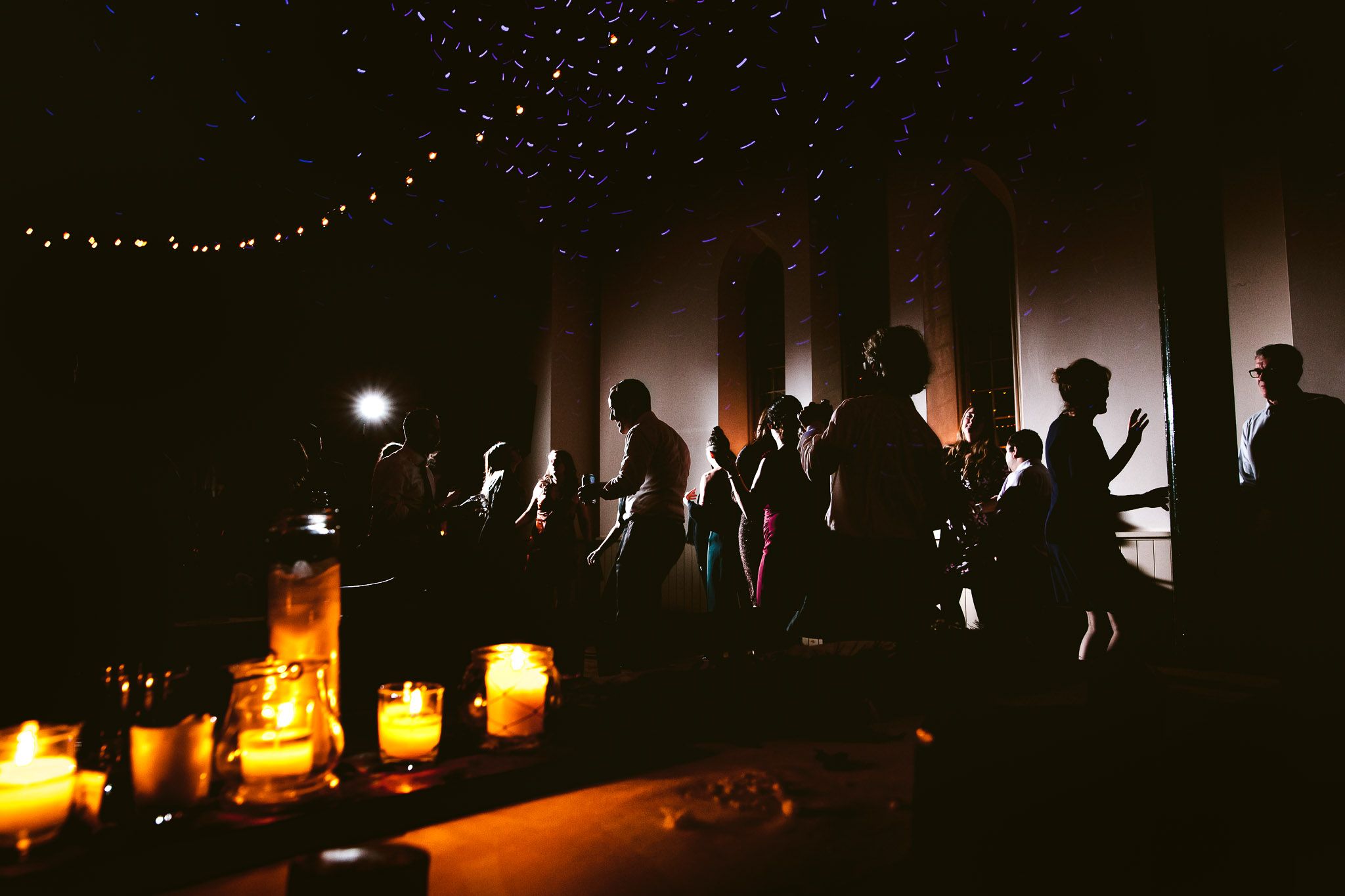 Silhouettes of guests dancing at reception with candlelight and string lights creating ambiance.
