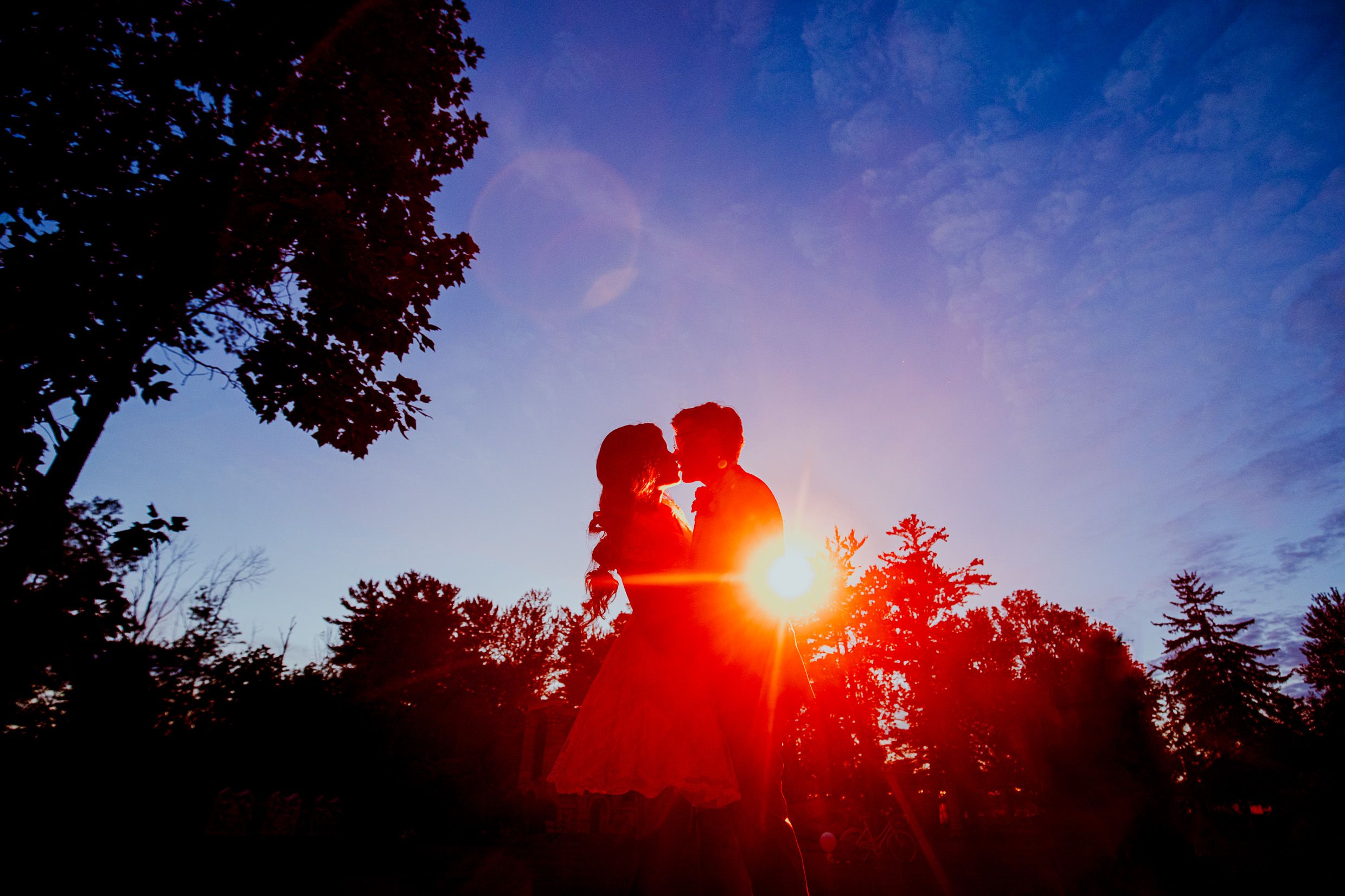 Silhouetted couple embraces against dramatic sunset sky with trees in background.