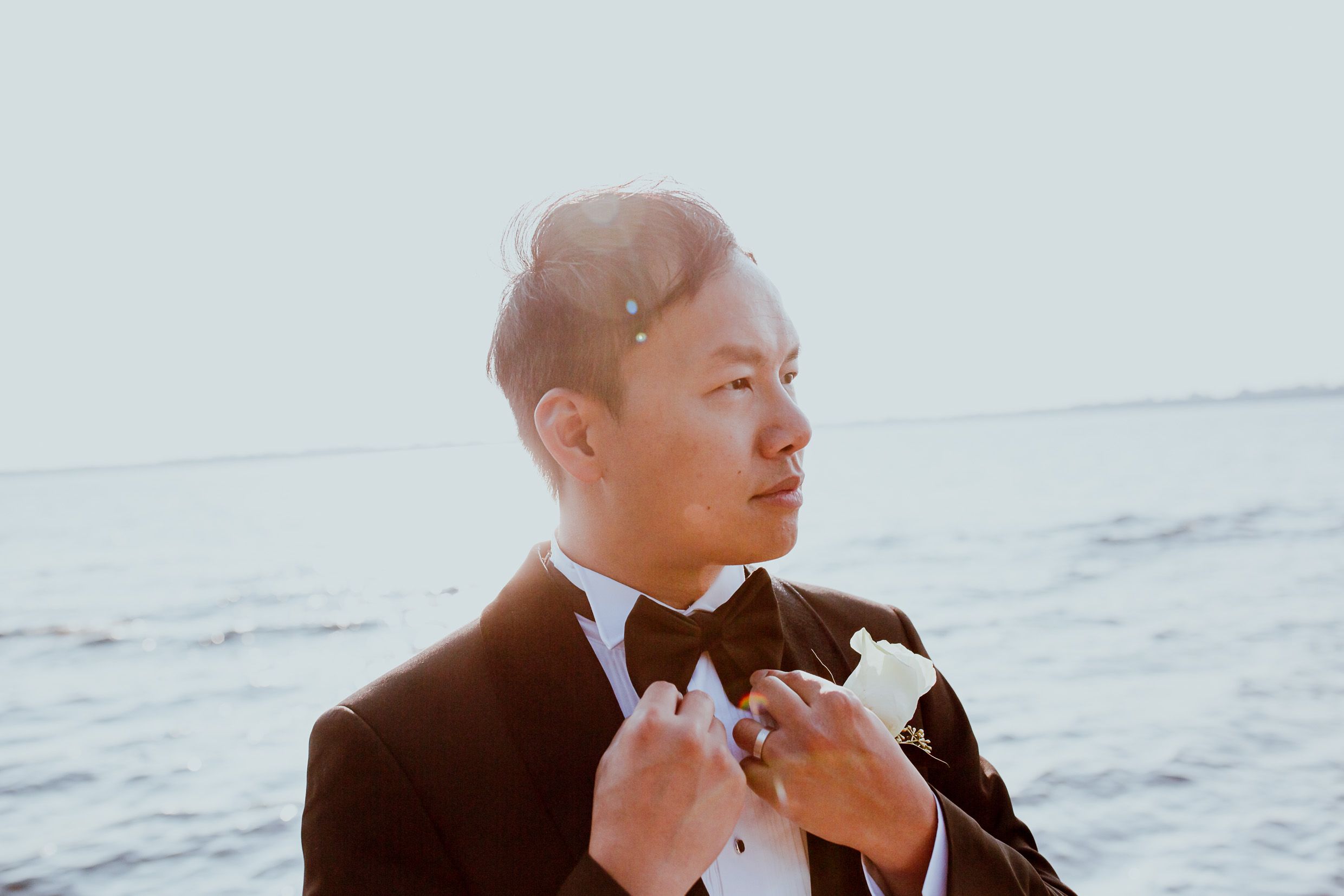 Groom adjusts bowtie while standing near ocean at sunset.