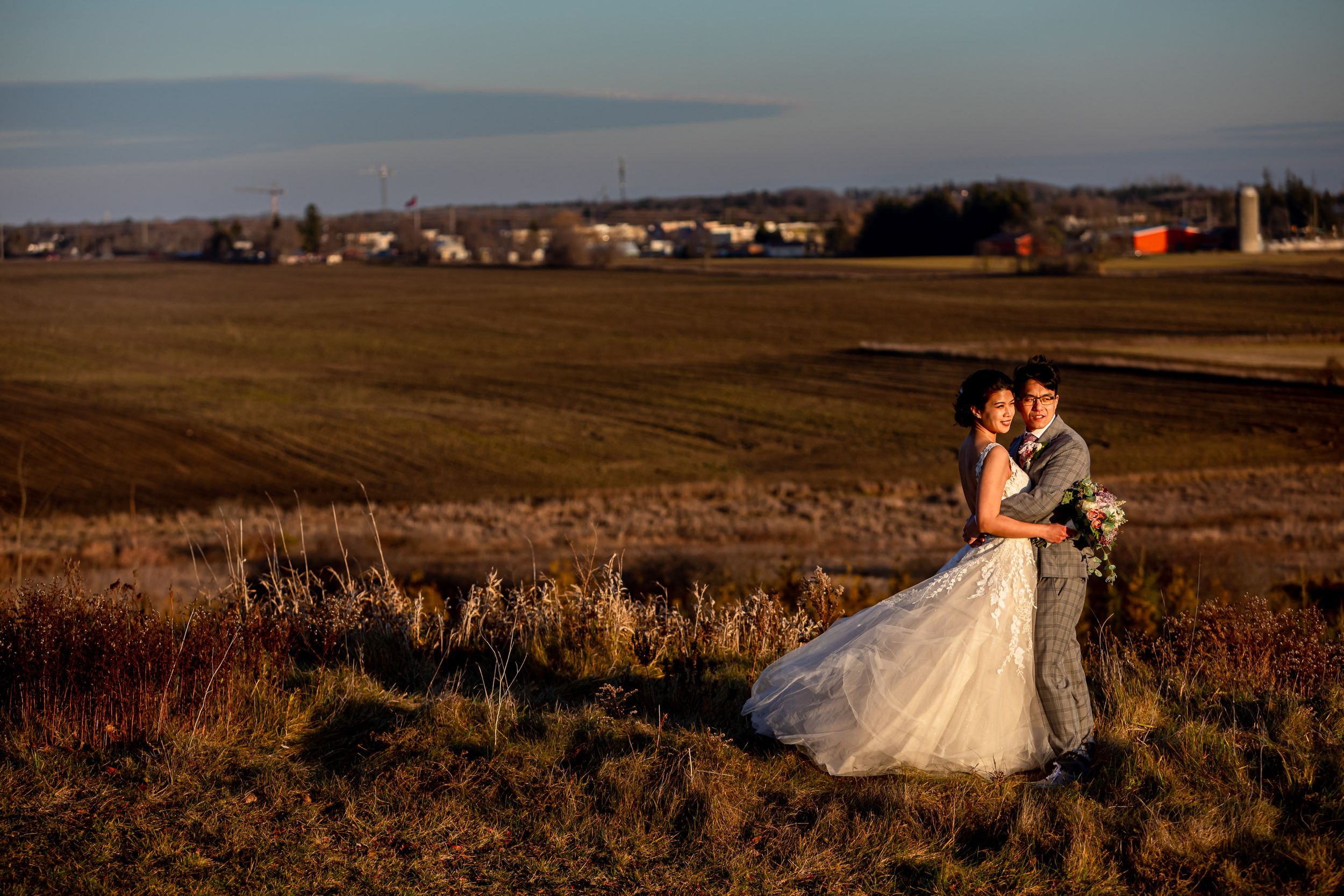 Newlyweds embracing in an open field during golden hour with houses in the background.