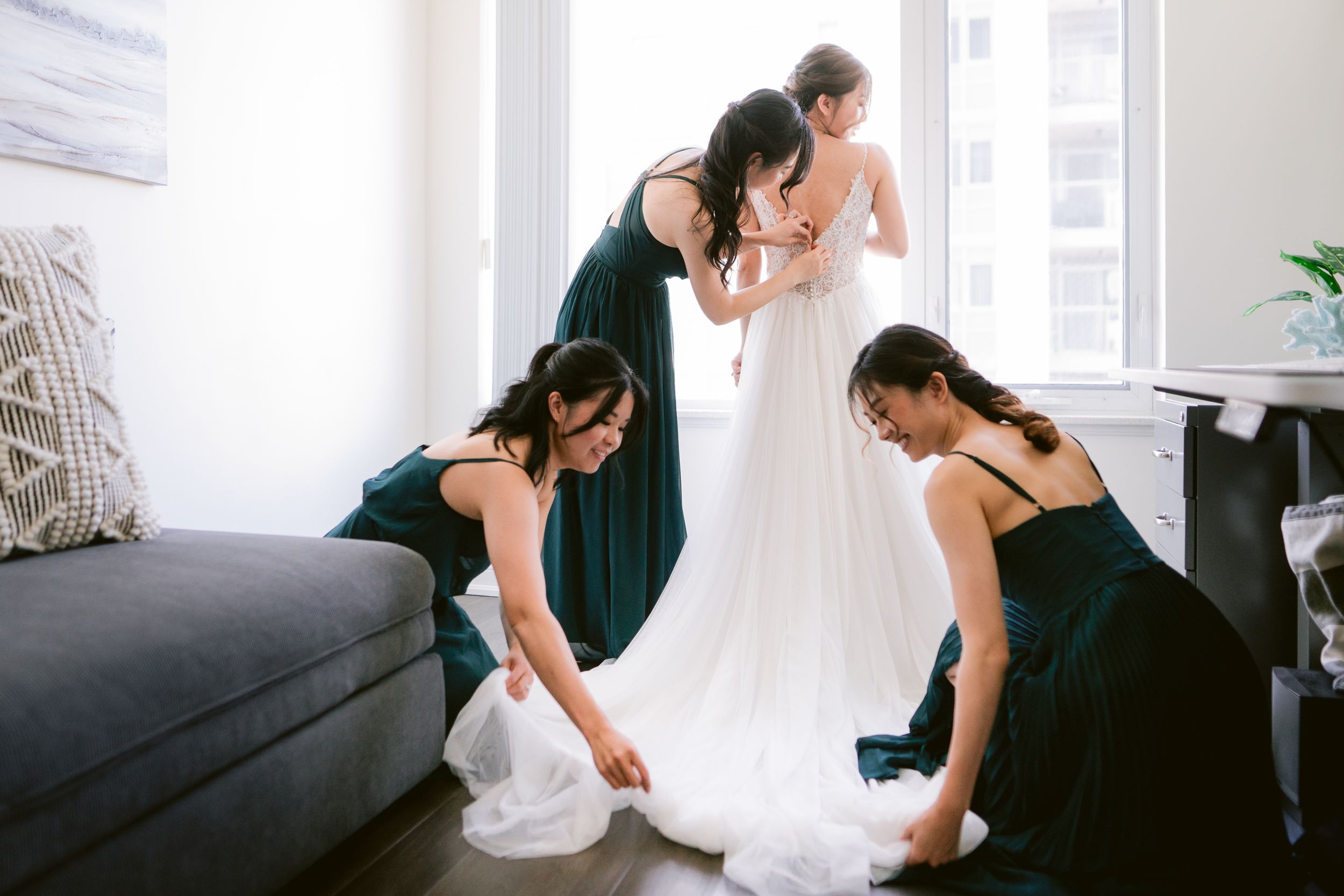 Bridesmaids in dark dresses help adjust wedding gown in bright modern room.