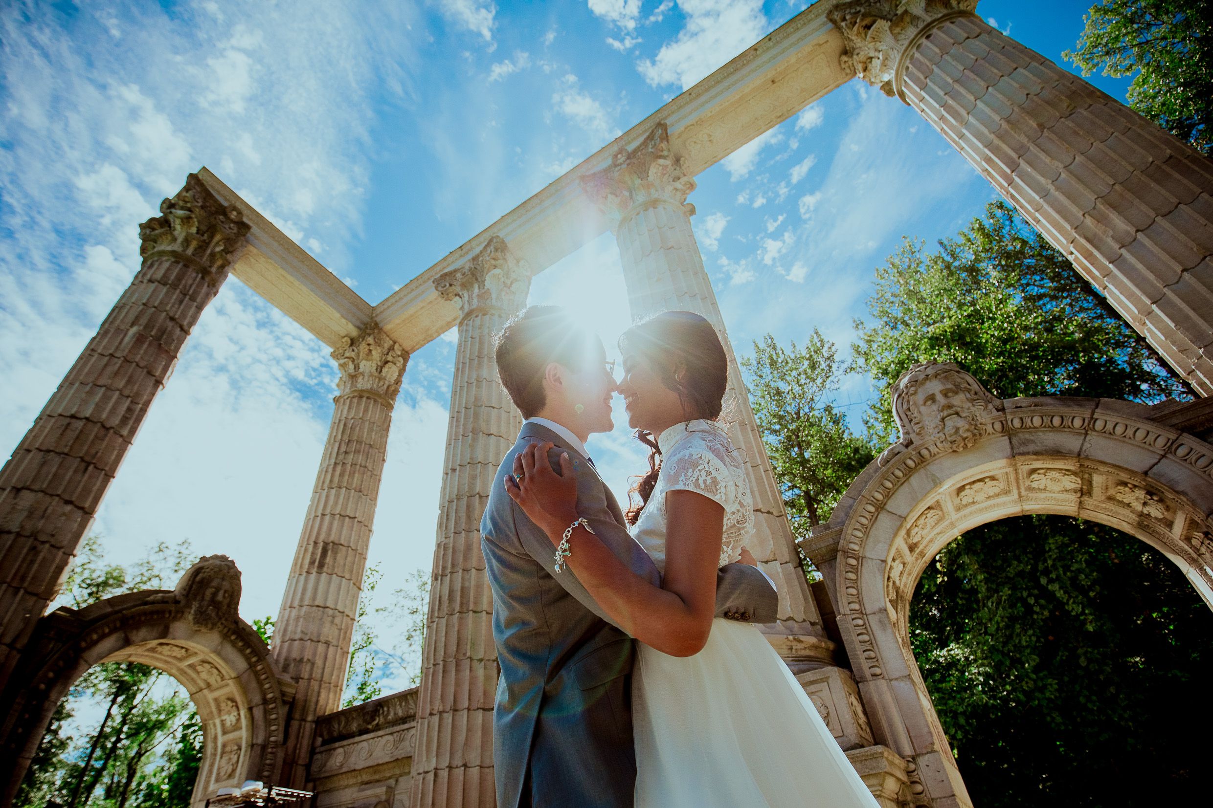 Couple embraces under sunlit stone columns and archways in romantic outdoor setting.