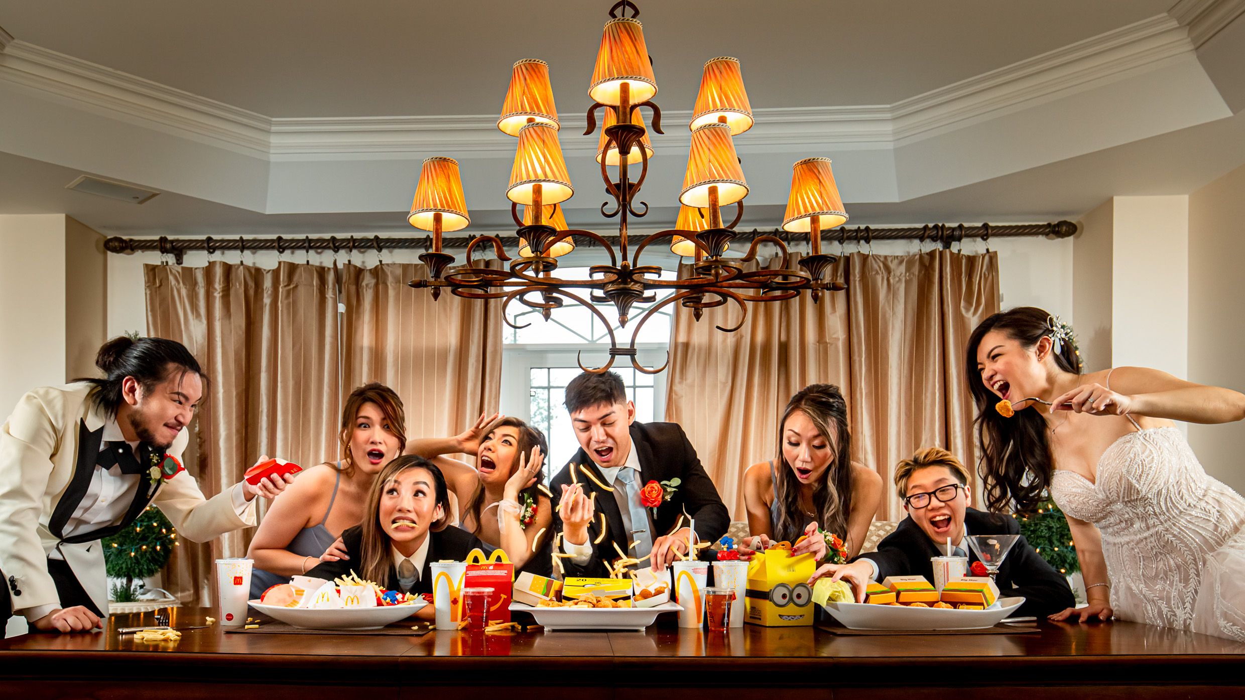 Wedding party gathered around dining table having animated conversation under warm chandelier lighting.