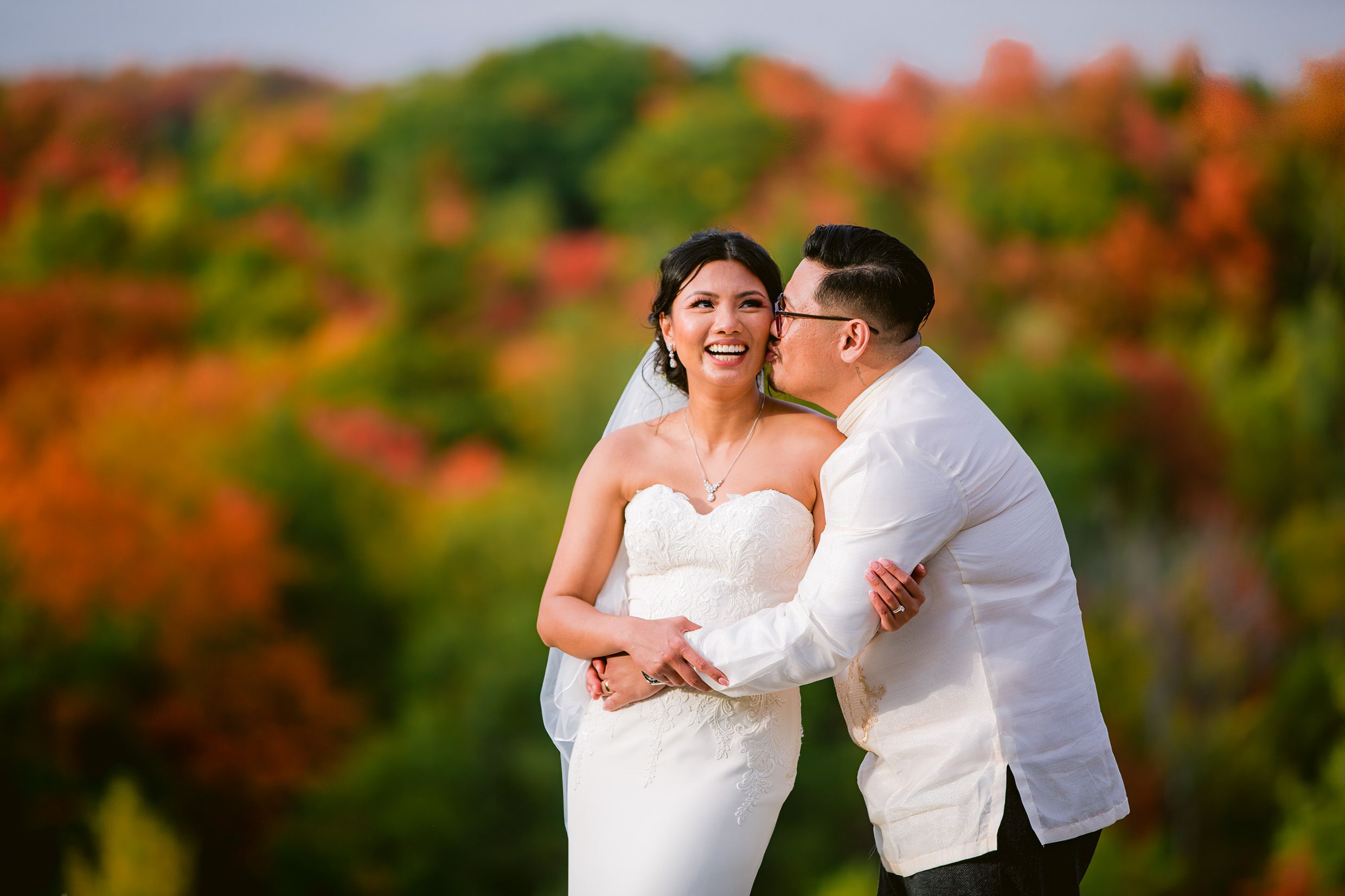 Newlyweds share a joyful moment against a backdrop of autumn trees.