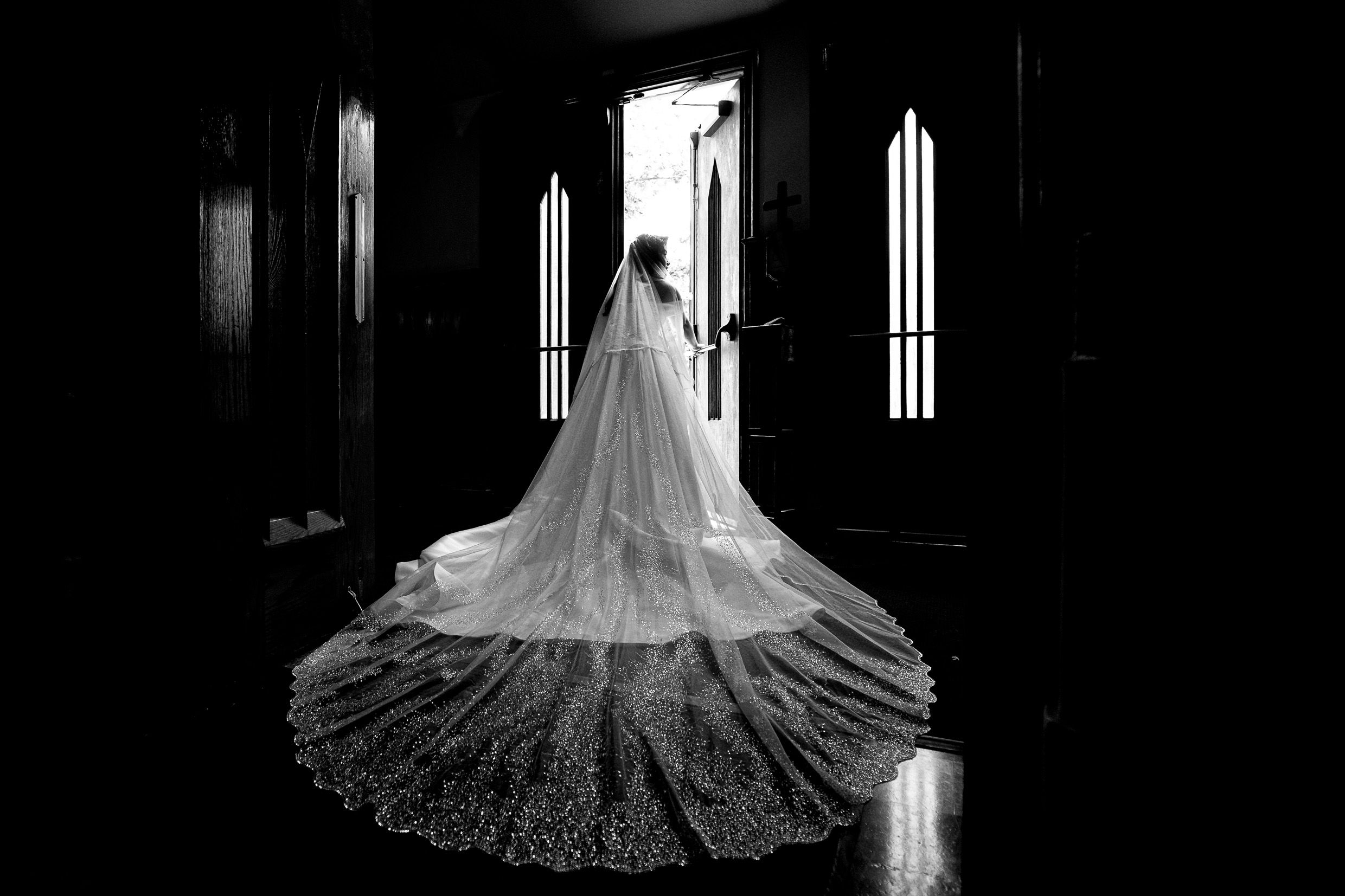 Bride in flowing lace gown stands in dramatic lighting by church windows.