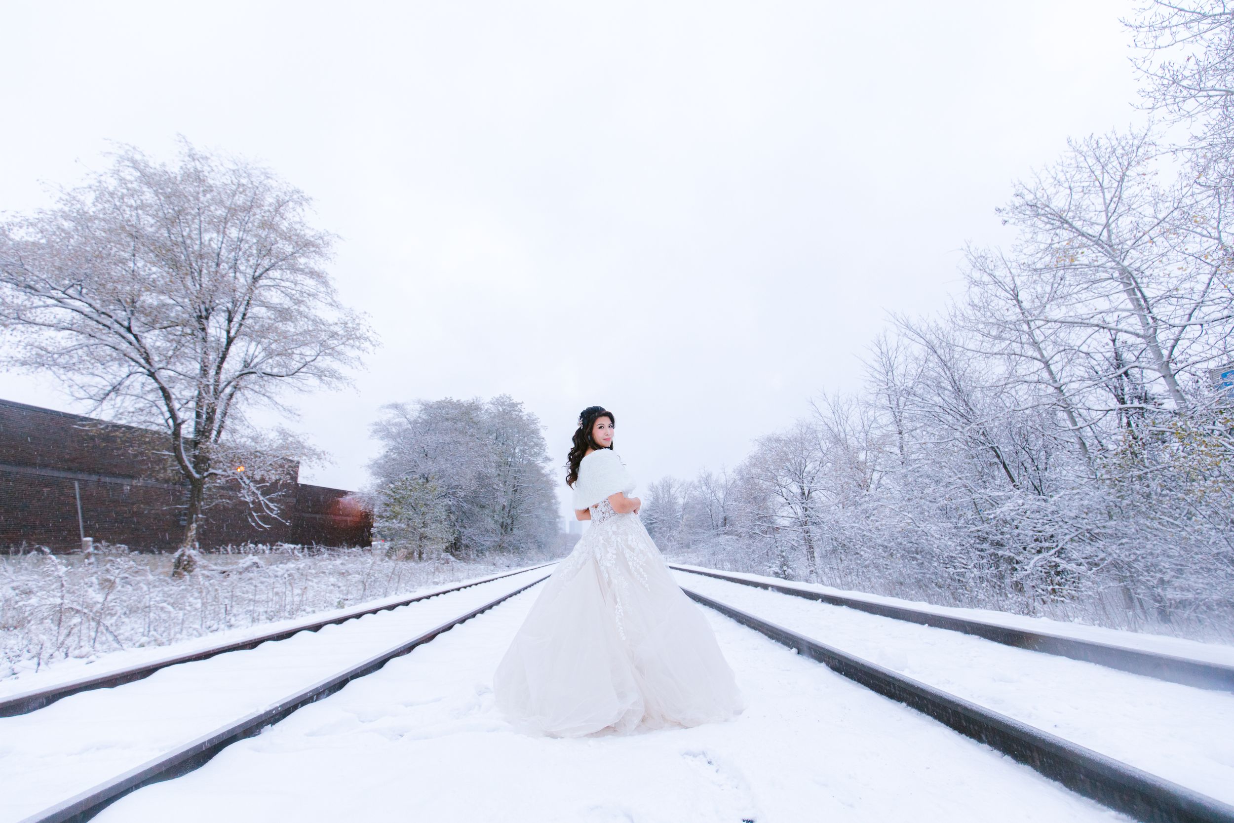 A bride in a white dress poses on snow-covered train tracks surrounded by winter scenery.