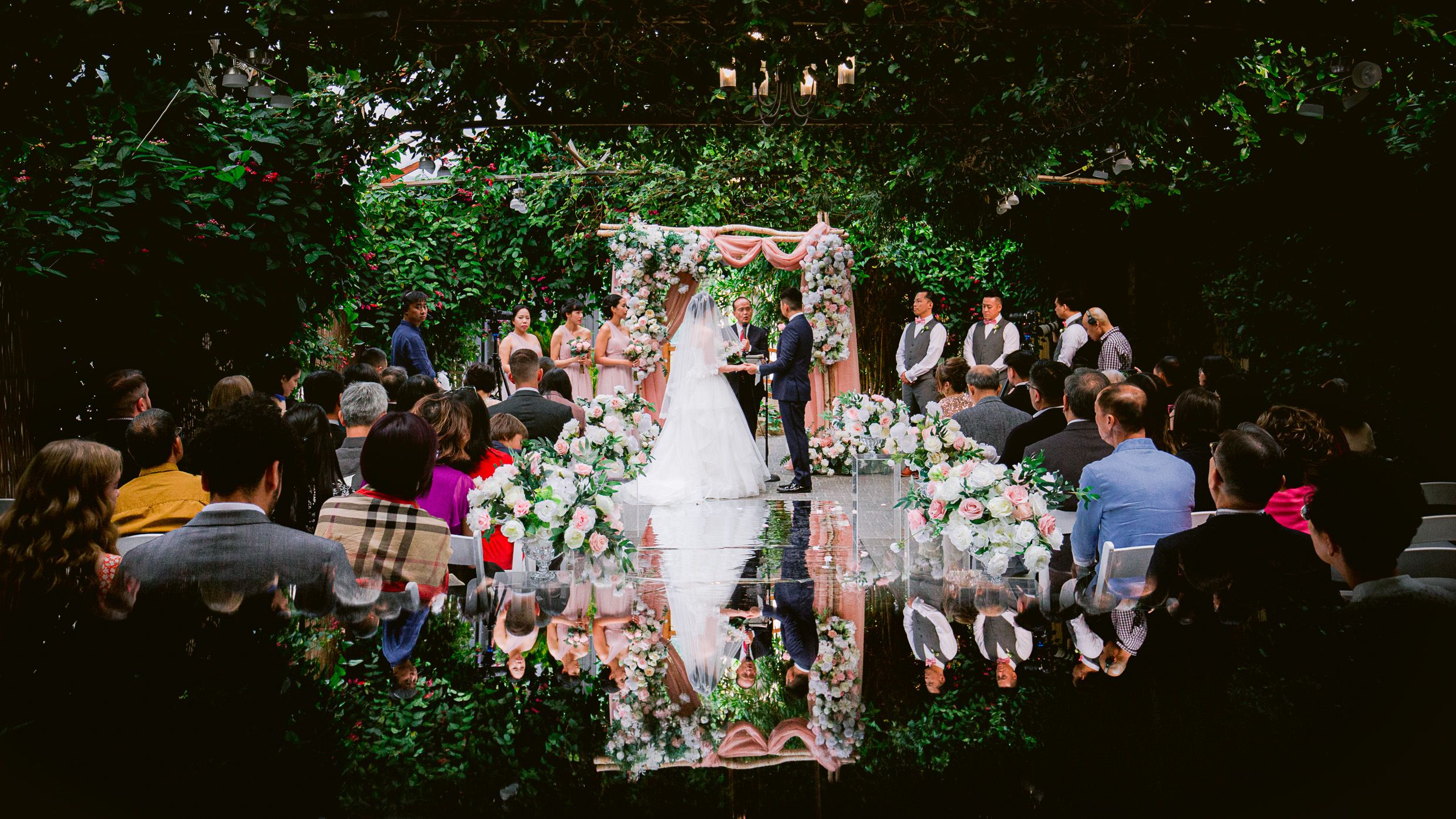 Garden wedding ceremony with floral archway and seated guests viewed from above with reflection.