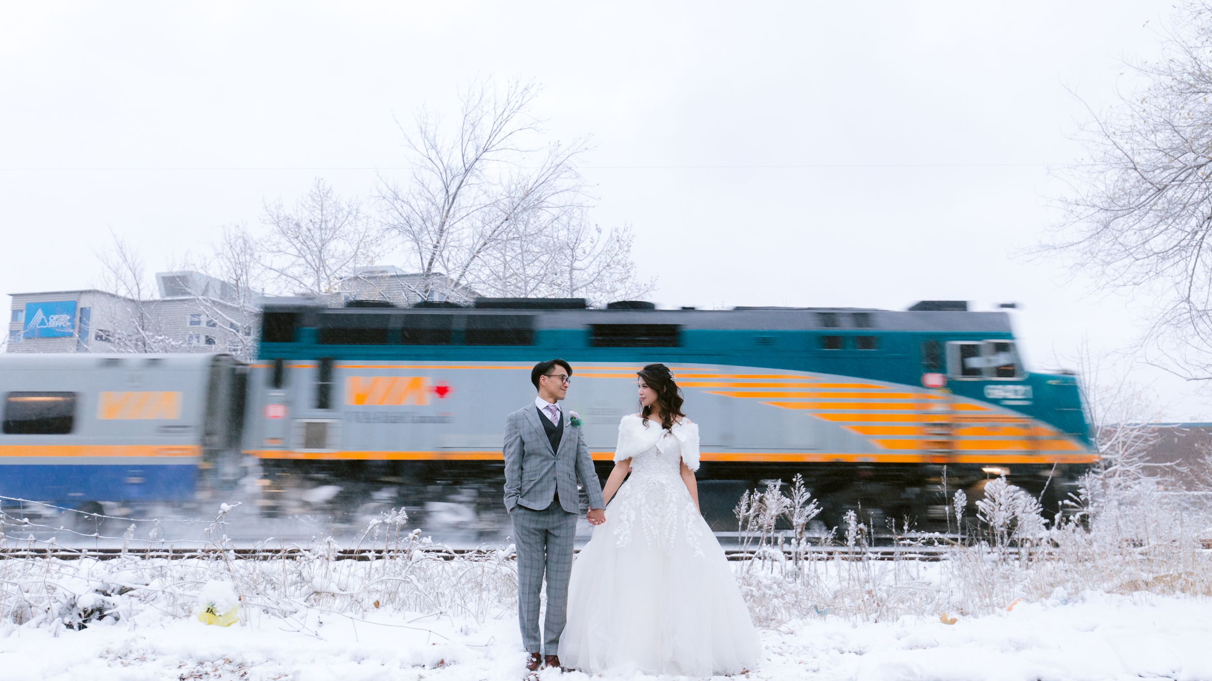 A wedding couple poses in front of a passing train on snowy railroad tracks.