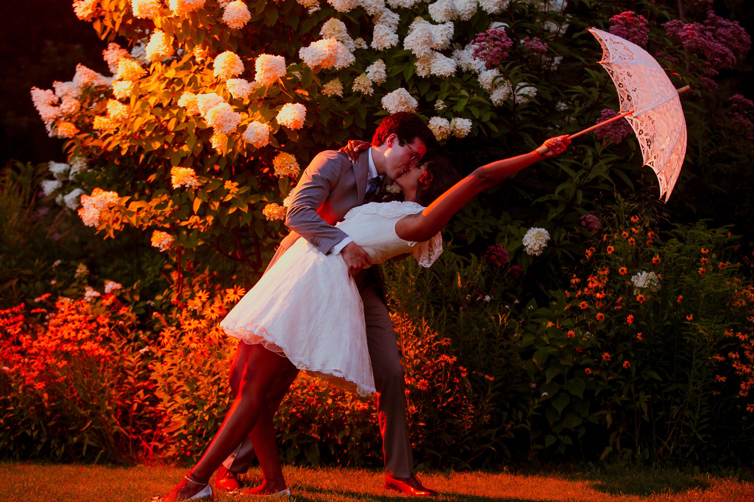 Couple dances with red umbrella in garden with glowing lights at dusk.