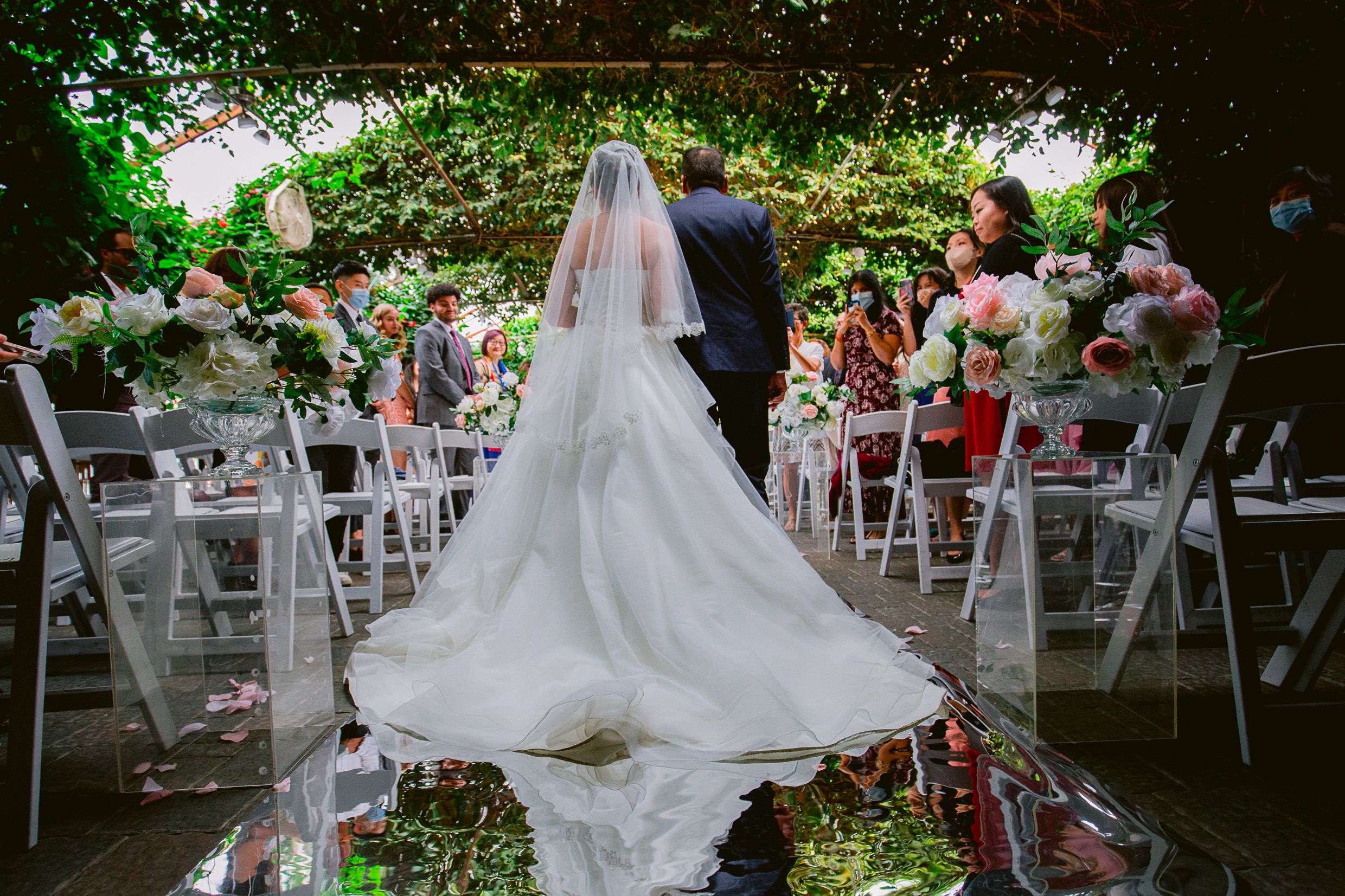 Wedding ceremony in a garden setting with white chairs and floral arrangements reflected in a puddle.