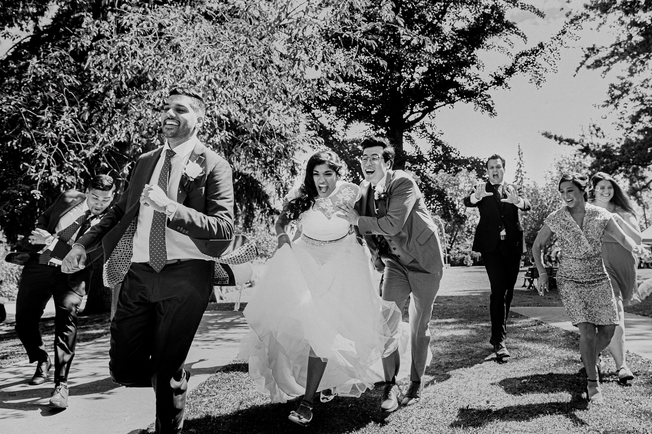 Wedding party joyfully runs together down a path in black and white photography.