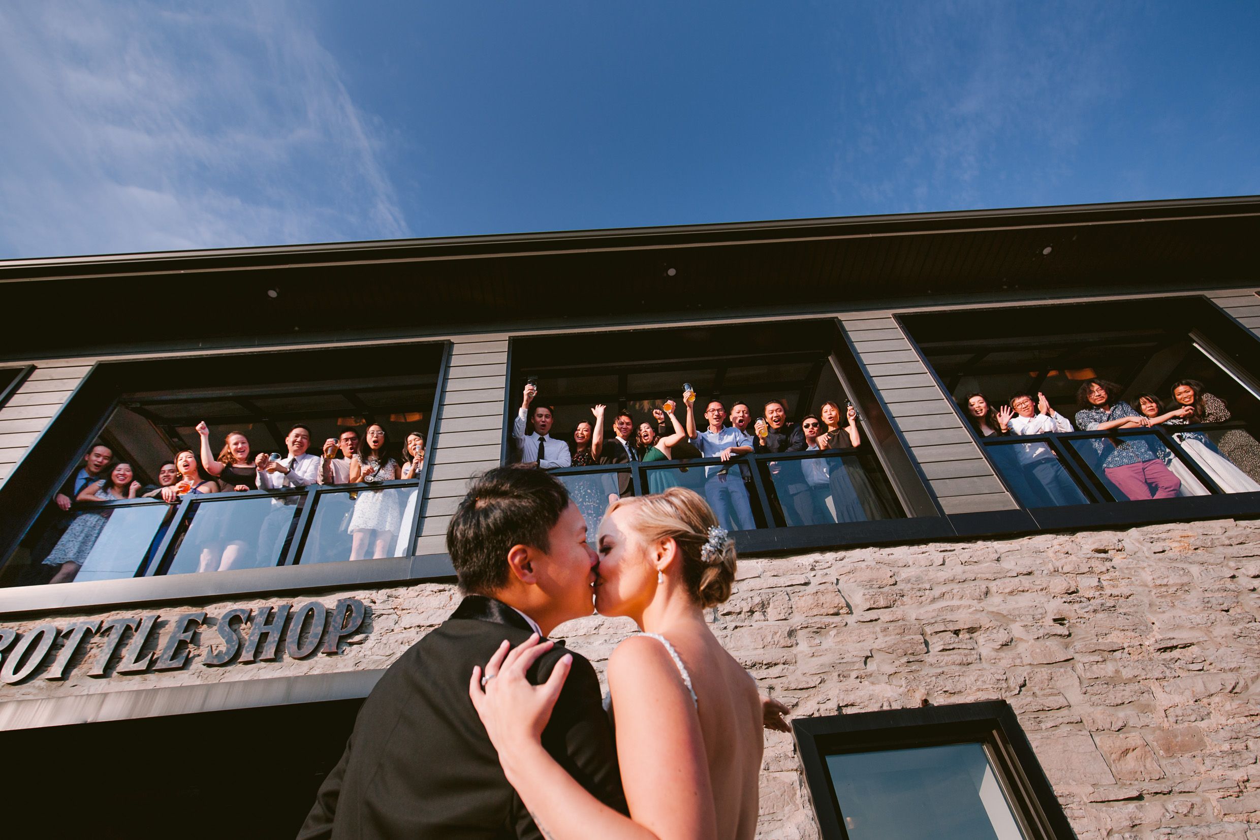 Wedding guests cheer from balcony of brick building as couple shares romantic moment below.