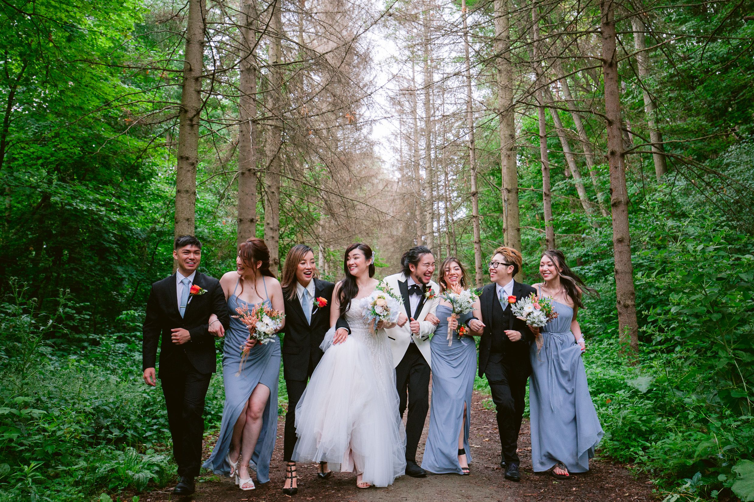 Wedding party poses together on forest path surrounded by tall pine trees.