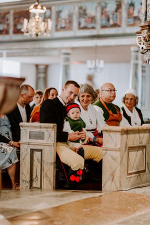 Wedding guests seated in ornate church pews with chandelier and decorative details overhead.