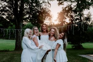 A joyful group in white dresses posing together at sunset with string lights.