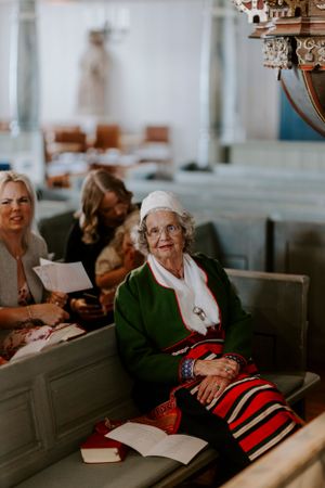Elderly guest in green cardigan sits in church pew with striped skirt and book during wedding ceremony.