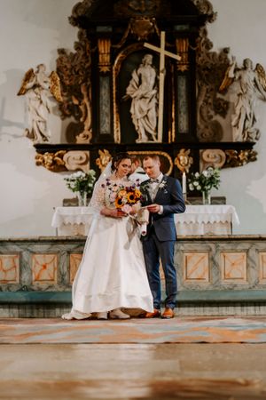 Bride and groom stand together before ornate baroque church altar with religious statues and cross.