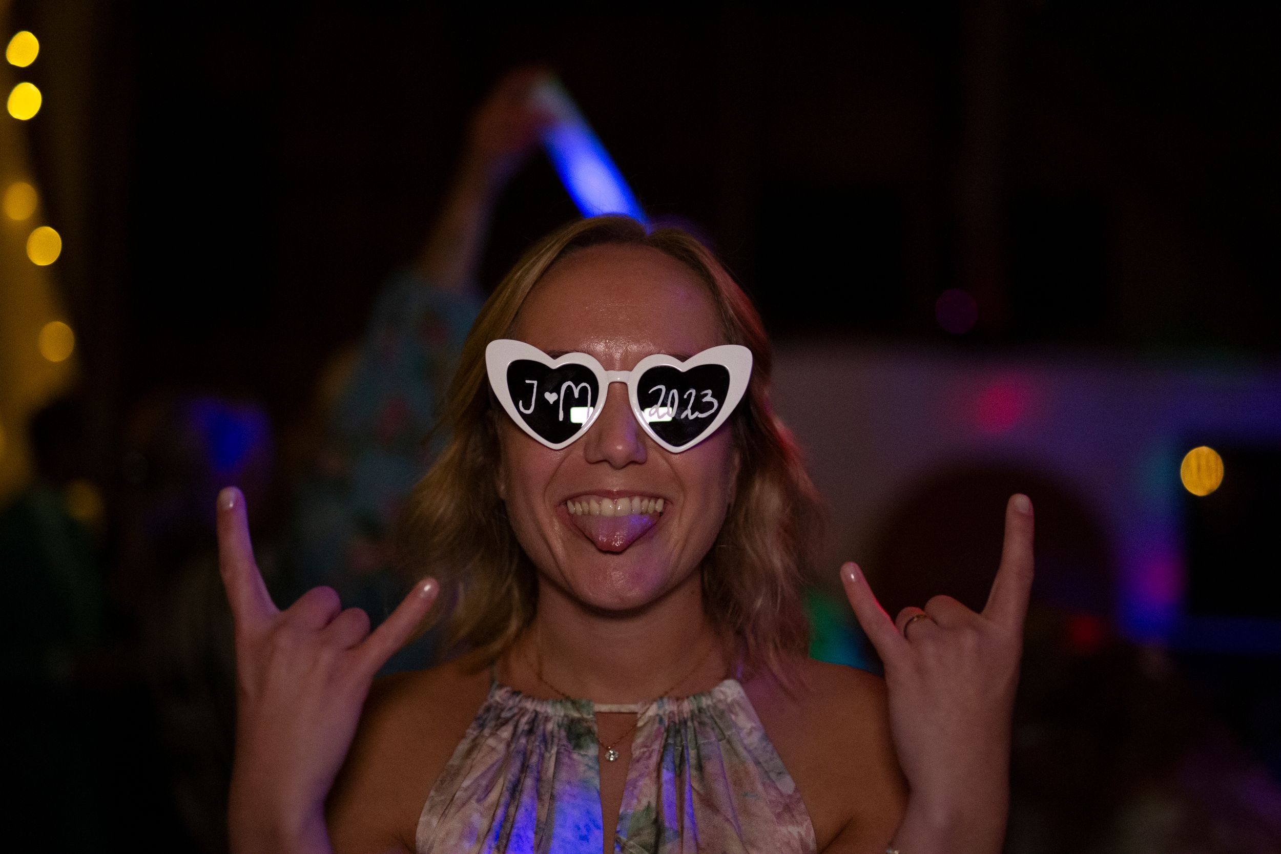 Party guest wearing heart sunglasses makes rock gesture against dark background.