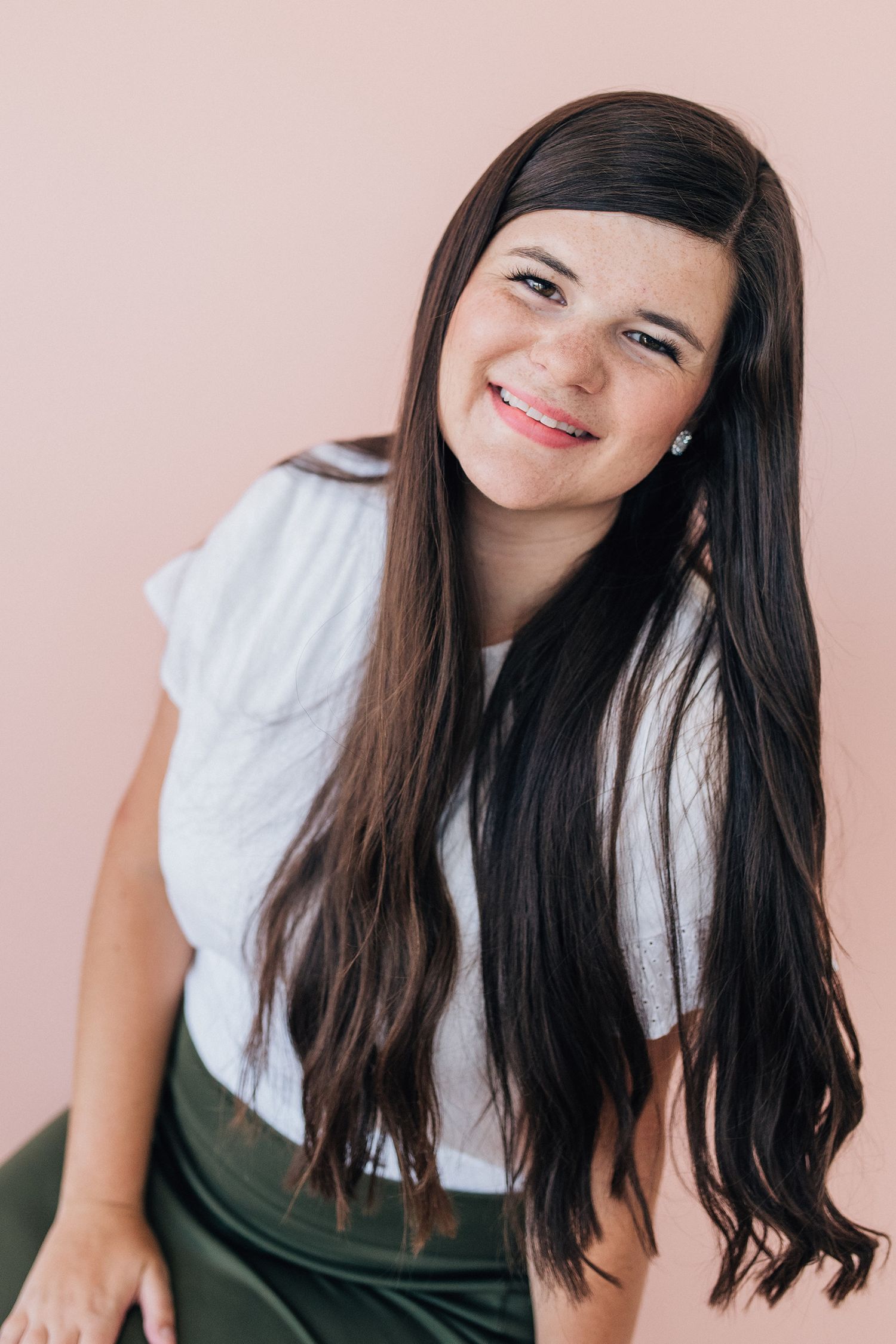 Portrait of a smiling young woman with long dark hair wearing a white top against a pink background.