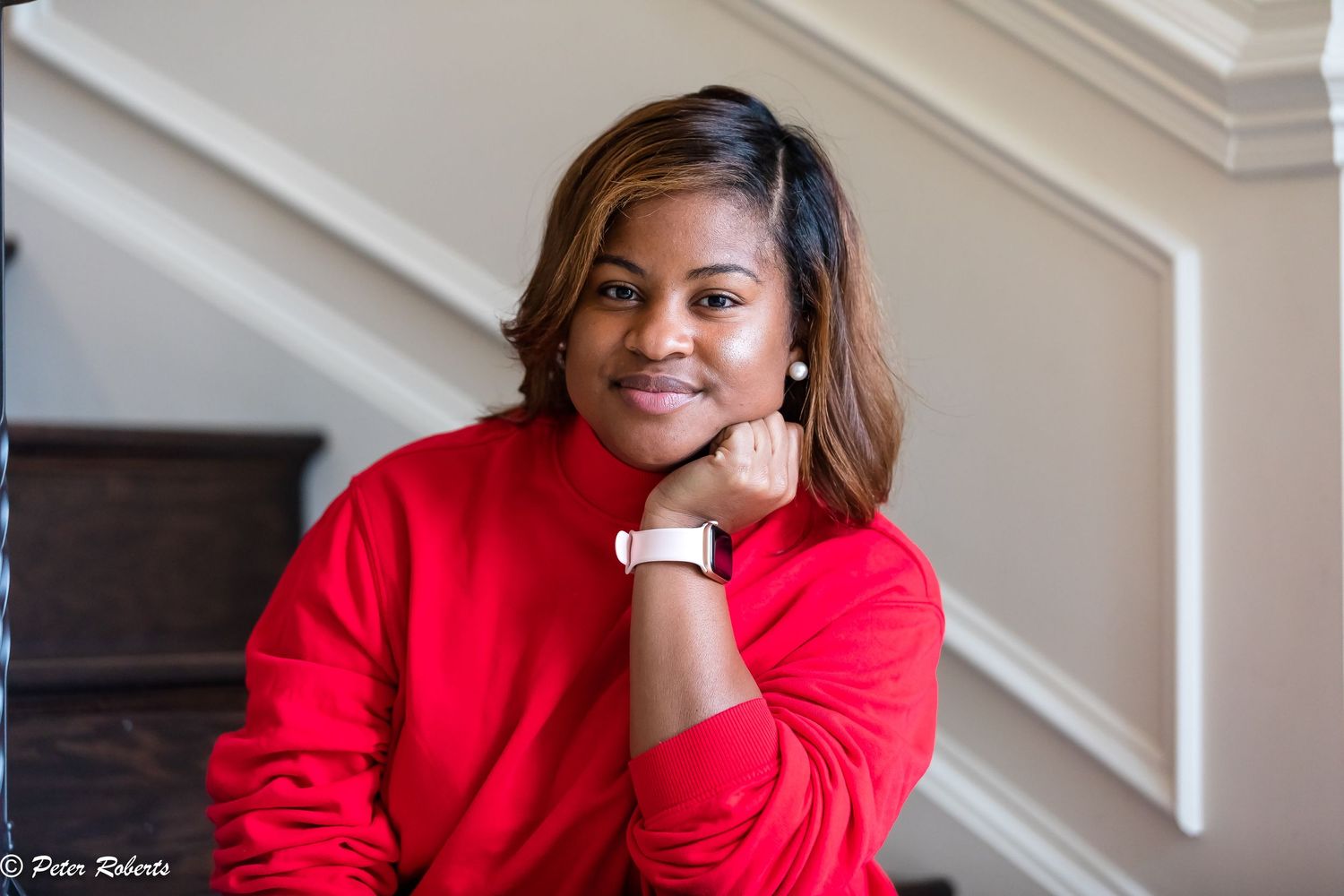 Smiling young woman in red sweater posing on staircase with chin resting on hand.