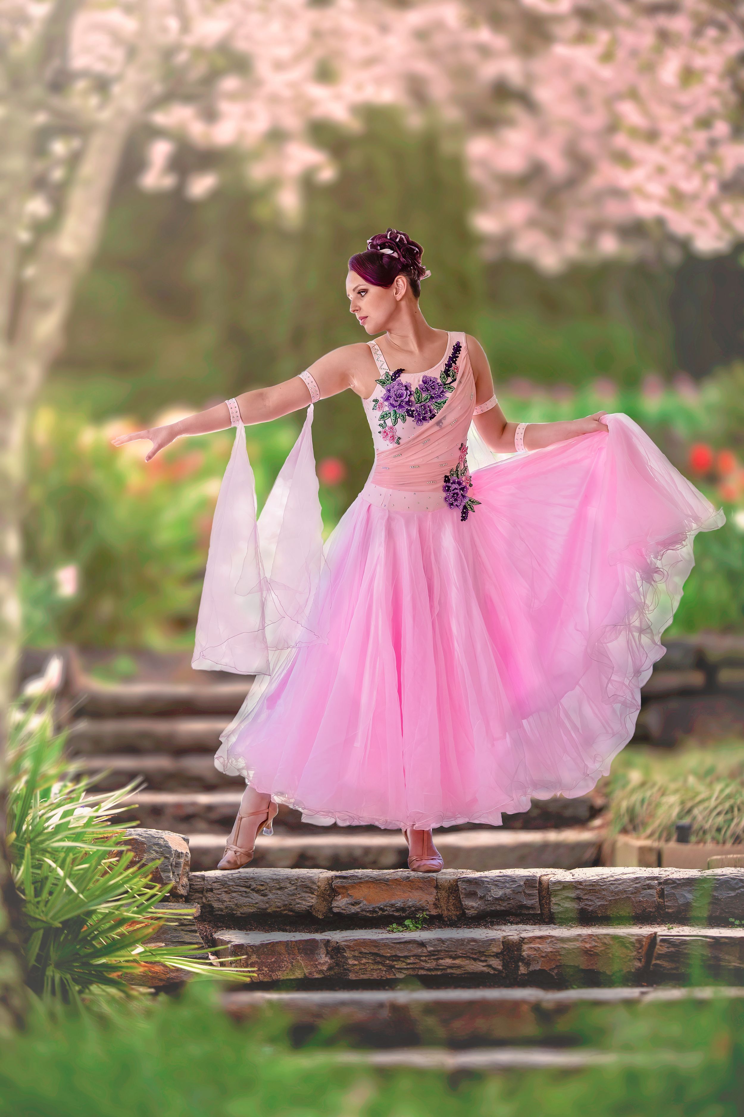 Woman wearing a pink ballroom dancing gown striking a dance pose on stone steps in beautiful garden.