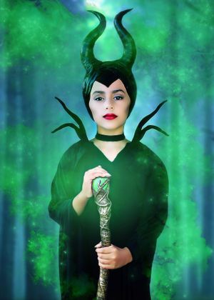 Child dressed as Disney character Maleficent surrounded by green mist holding a glowing staff in a menacing forest.