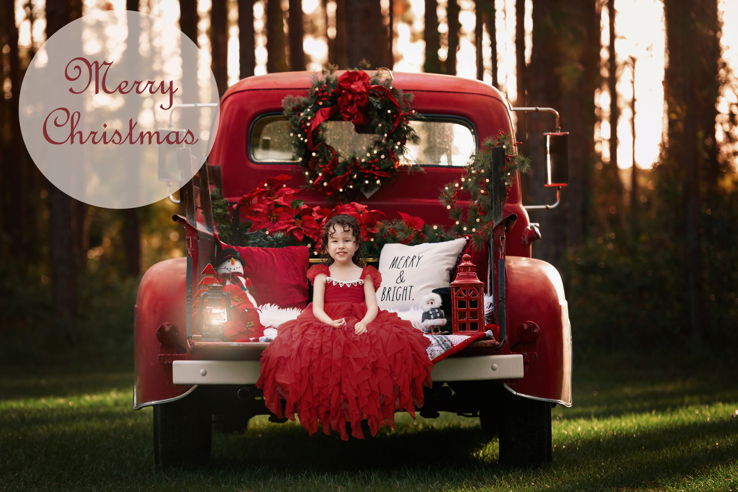 Little girl in red gown sits on tailgate of an old American truck with the rear tray filled with Christmas decorations.