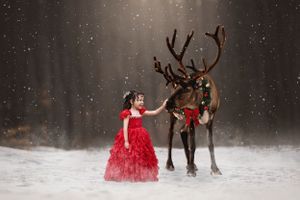Little girl in red gown standing in snow in front of a forest petting a reindeer wearing a festive wreath as a collar.