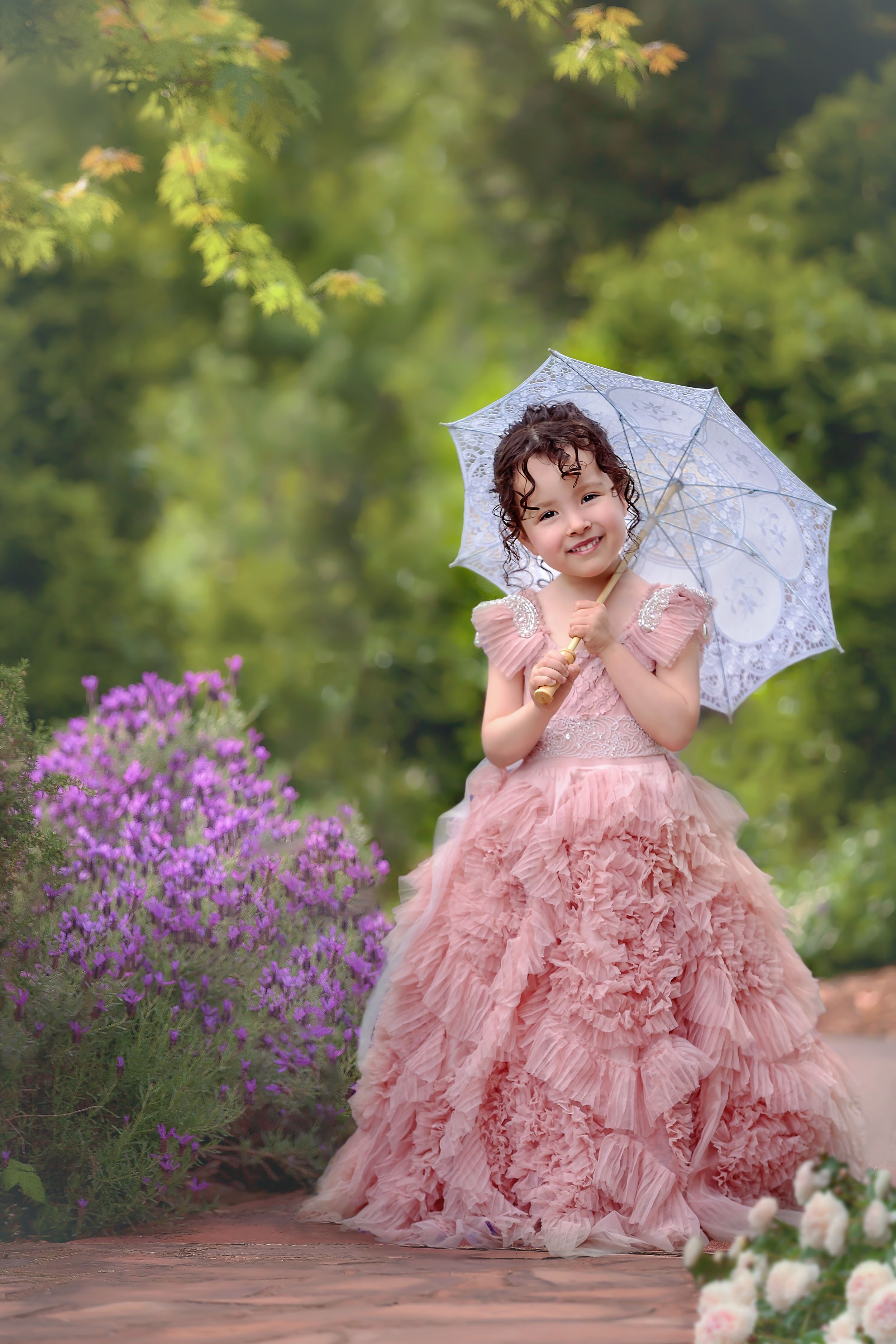 Small girl in a long ruffled pink gown holding an open lace parasol standing in a beautiful garden.