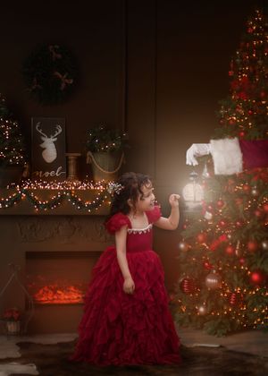 Little girl in red gown standing in front of a Christmas scene looking at a lantern held by Santa who is out of frame.