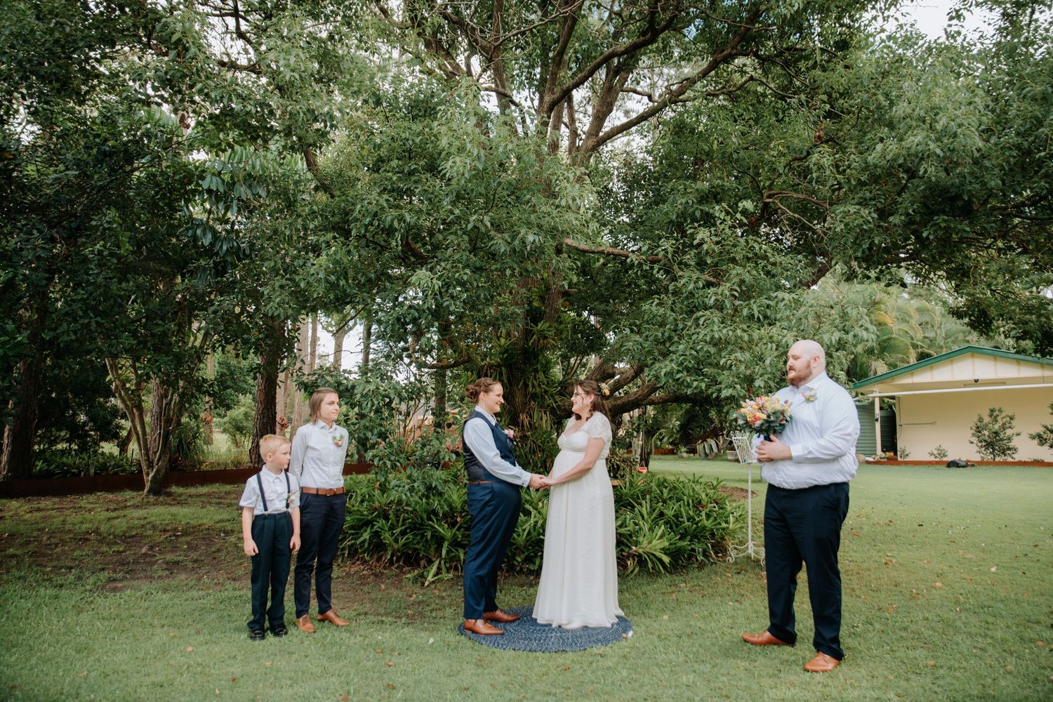 An outdoor wedding ceremony taking place under green trees with a small group gathered.