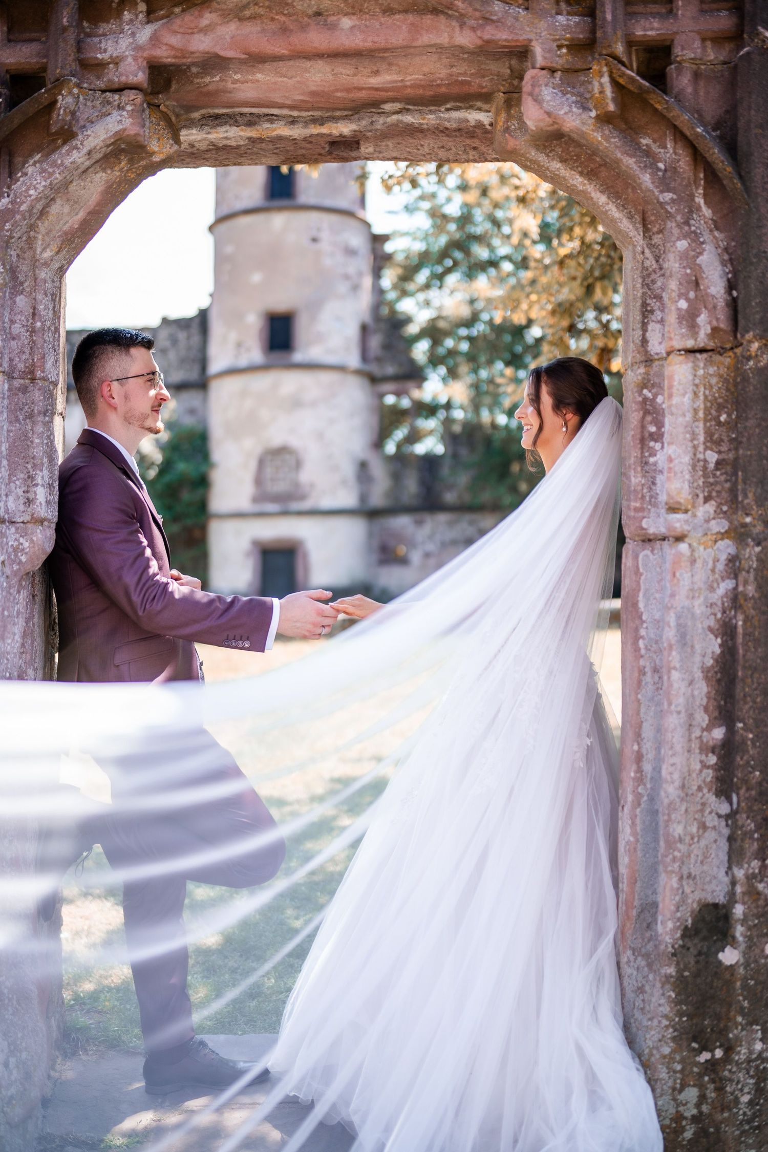 A wedding couple poses under a stone archway with a castle tower in the background.