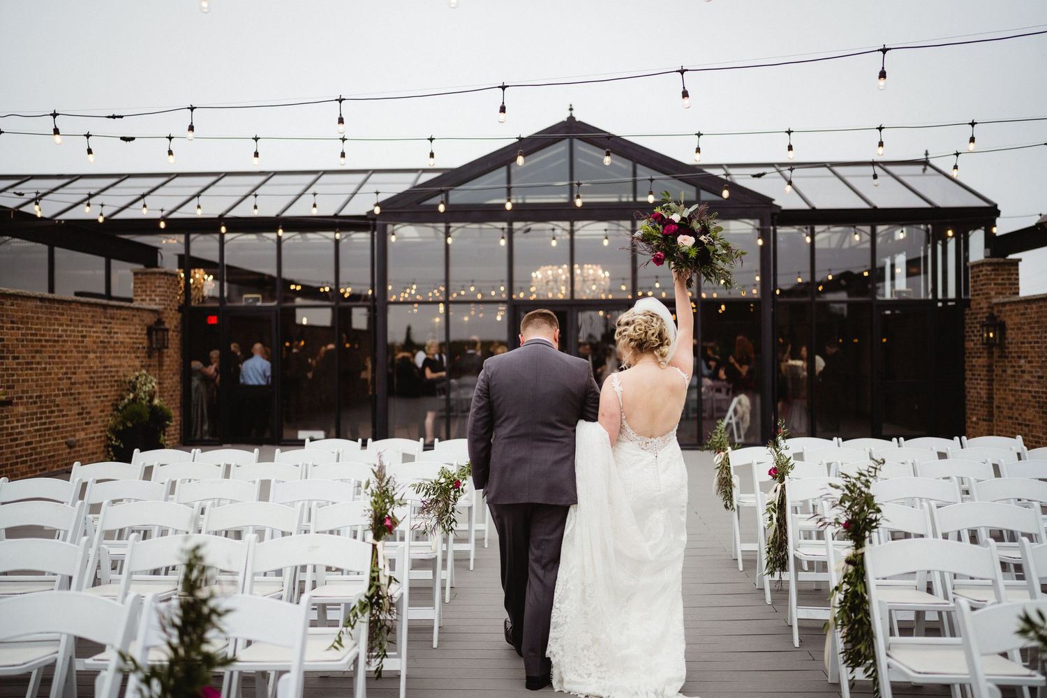 Father walks bride down aisle at outdoor ceremony with greenhouse and string lights backdrop.