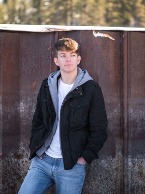 Young person in a black jacket and gray hoodie poses against rustic metal wall in outdoor portrait setting.