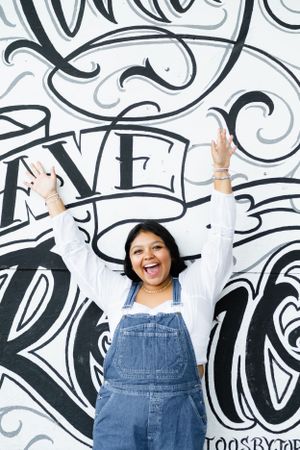 A joyful person in denim overalls raises their arms against a wall with decorative black and white typography artwork.