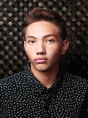 A high school senior portrait against dark patterned background wearing black and white patterned collared shirt.