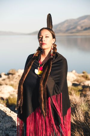 A high school senior in traditional Native American dress with feathers and fringe stands against a serene lakeside mountain backdrop.