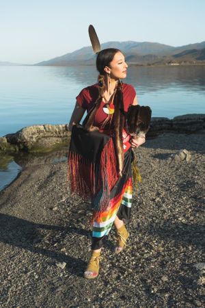 A high school senior in Indigenous wear in ceremonial regalia stands gracefully by a serene lake with mountains in the background.