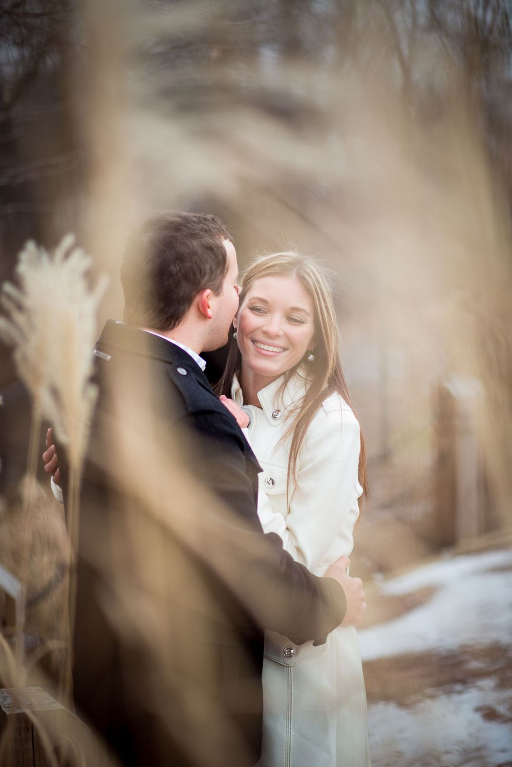 A recently engaged couple embracing at the Naperville Riverwalk with wildgrass plumes and snow in the background.