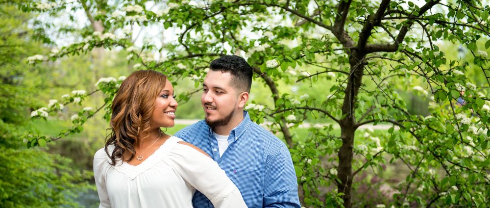 A recently engaged couple smiling at eachother with trees in the background at the Naperville riverwalk.