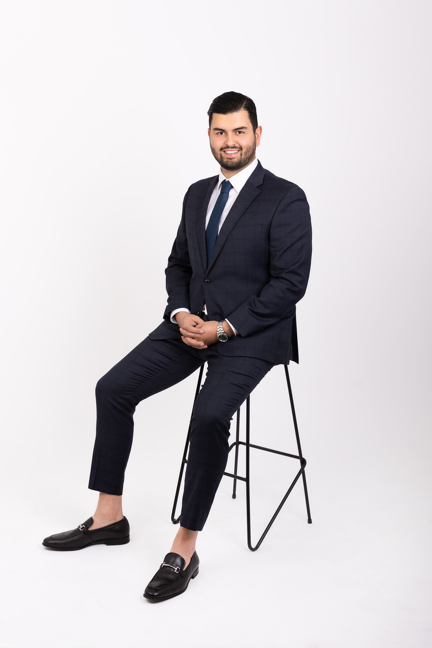Smiling businessman in navy suit and tie sits on a stool with hands clasped in white studio.