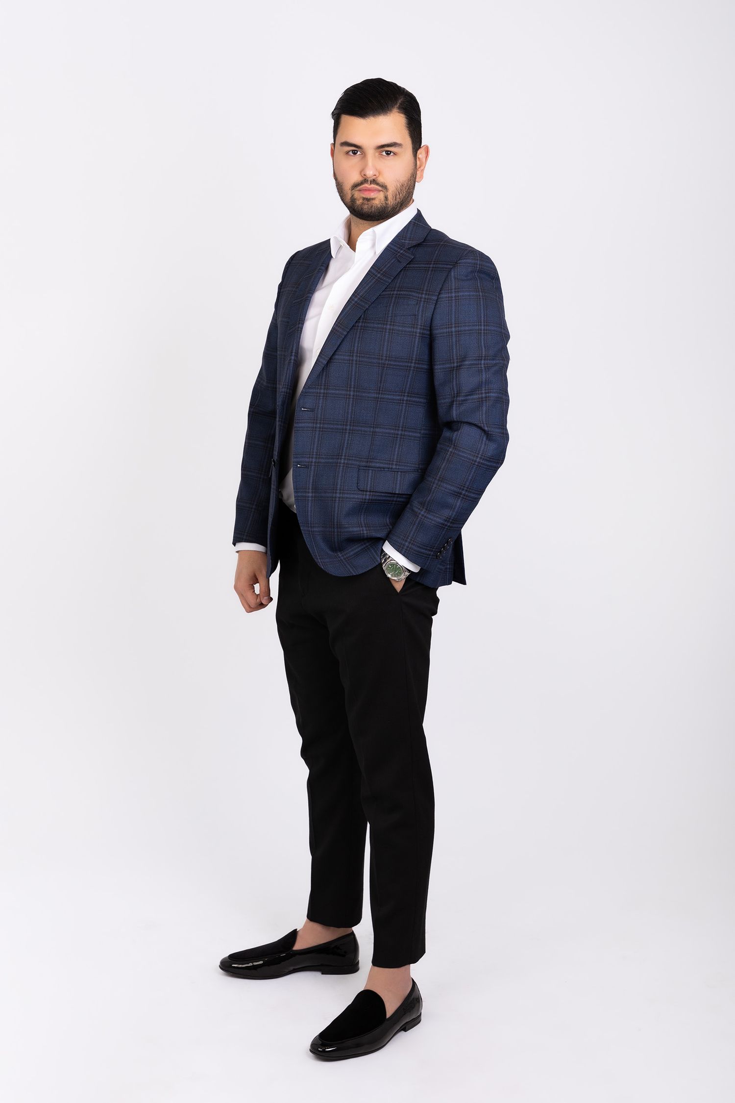Man in navy plaid blazer and black trousers poses confidently against a white studio background.